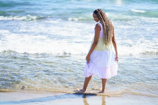 Black female walking along the shore of the beach wearing a beautiful long dress. Young girl enjoying her vacations.