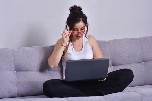 Beautiful brunette woman in red glasses on sofa using laptop