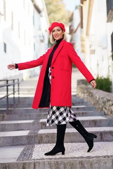 Happy blond woman in trendy coat with dress and beret with boots looking at camera while walking on street steps
