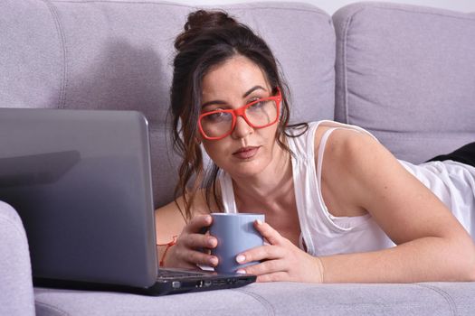 Beautiful brunette woman in red glasses on sofa using laptop