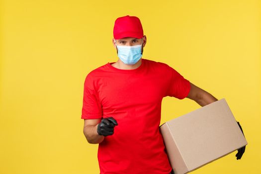 Express delivery during pandemic, covid-19, safe shipping, online shopping concept. Determined hardworking courier in red uniform and medical mask, holding package box and pointing finger camera.