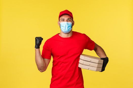 Food delivery, tracking orders, covid-19 and self-quarantine concept. Rejoicing smiling courier in medical mask and red uniform, holding pizza, bring order, fist pump in joy, success or achievement.