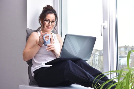 Home quarantine, protection from coronavirus. Caucasian brunette woman sit on window with computer