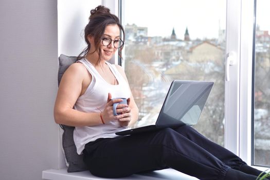 Home quarantine, protection from coronavirus. Caucasian brunette woman sit on window with computer