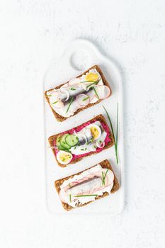 Savory smorrebrod, set of traditional Danish sandwiches. Black rye bread with anchovy, beetroot, radish, eggs, cream cheese on grey plate on a white stone table, top view, vertical