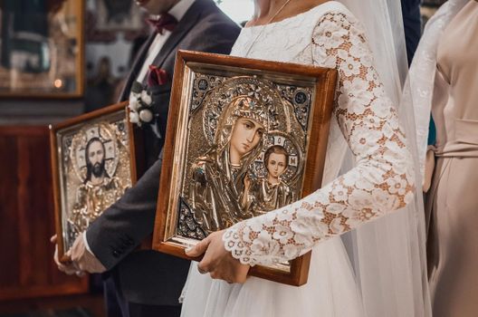 The bride and groom in a white dress are holding large icons of the Mother of God.