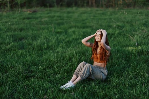 A red-haired woman with long, windswept hair sits outdoors on the grass in the park and smiles, the sunset light illuminating her face. The concept of harmony with nature outdoors. High quality photo