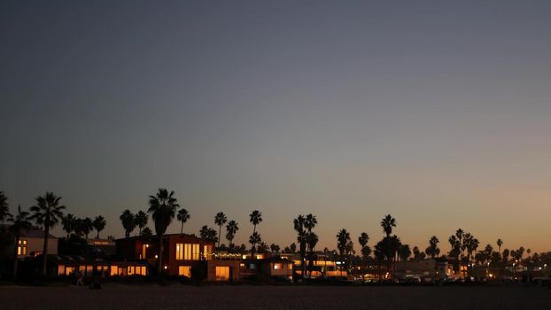 Palm trees silhouettes in twilight sky, California beach, USA. Beachfront palmtrees on summer coast, pacific ocean shore in dusk. Windows of houses or homes in evening. Dark contrast architecture.