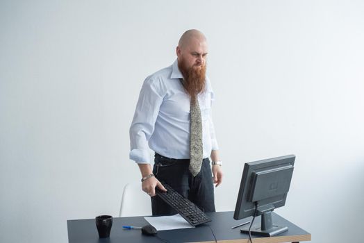 Angry bald man with a red beard in the office in a business suit crashes a computer. Manager with a nervous breakdown breaks the keyboard on the monitor