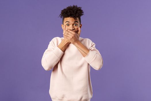 Portrait of shocked impressed young hispanic man found out gossip, heard rumour, trying not laugh shut his mouth with hands and express amazement and surprised with gaze, purple background.