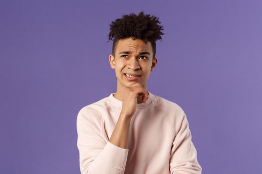 Close-up portrait of indecisive, puzzled young man facing difficult choice, look up thoughtful, touch chin and grimacing while thinking, making-up idea or decision, purple background.
