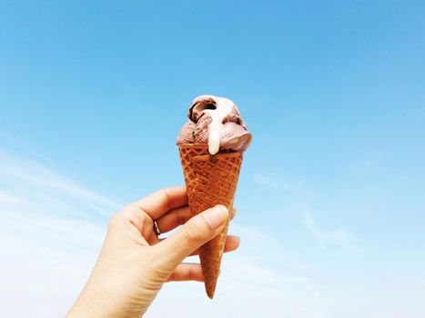 Woman's hand holding wafer cone of chocolate ice cream against bright blue sky background on a sunny day
