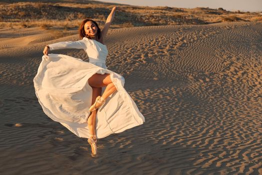 A girl in a fly white dress dances and poses in the sand desert at sunset.