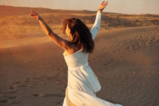 A girl in a fly white dress dances and poses in the sand desert at sunset.