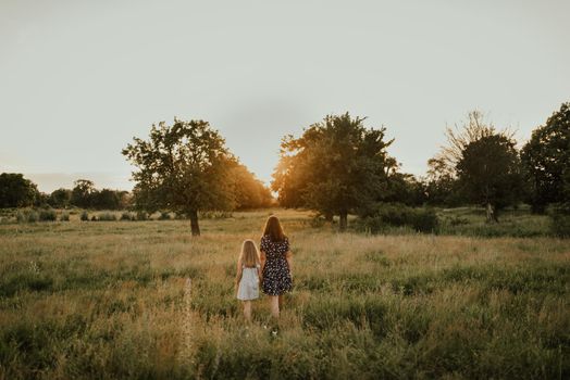 European family mom and daughter walk holding hands Along the tall green grass towards the sunset in summer. meadow and tall trees through which the rays of the sun make their way