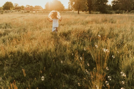 a little blonde girl dressed in light-colored clothes and a white T-shirt runs away from soap bubble. Along the tall green grass towards the setting sun. Warm summer sunny weather at sunset.
