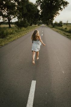 A small blonde European girl in a blue dress runs away along a clean asphalt road with markings where trees grow on the sides. Summer warm weather After sunset.