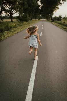 A small blonde European girl in a blue dress runs away along a clean asphalt road with markings where trees grow on the sides. Summer warm weather After sunset.