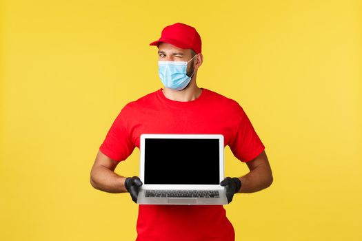 covid-19, safe shopping, express delivery and order tracking concept. Handsome courier in red uniform, medical mask and gloves, wink camera and showing page site on laptop screen.