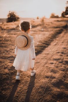 little girl in a white dress with curly hair goes towards the sun at sunset. boater hat. white socks with a pattern and sandals. The background is beautifully blurred. Soap bubbles fly in the air.