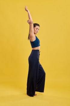 Side view of slim girl standing, raising hand, looking down, with closed eyes. Brunette with pony tail stretching, meditating, yoga pose practicing. Concept of new age and yoga.