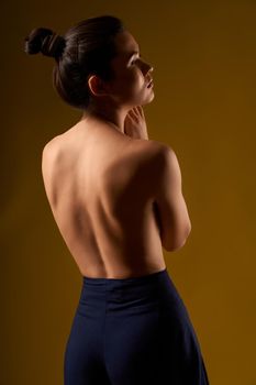 Back view of seductive female with bare back standing, looking forward. Brunette with hair bun doing yoga, breezing, meditating, relaxing. Isolated on yellow studio background.