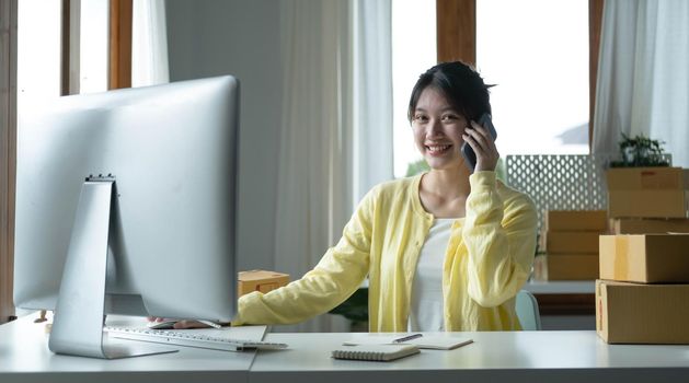 A portrait of Asian woman, e-commerce employee sitting in the office full of packages on the table using a laptop and smartphone, for SME business, e-commerce, technology and delivery business..