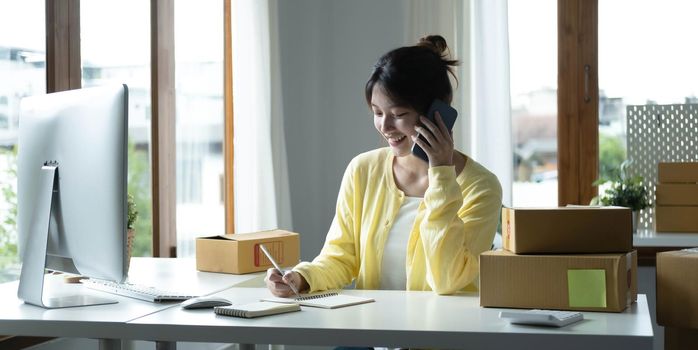 A portrait of Asian woman, e-commerce employee sitting in the office full of packages on the table using a laptop and smartphone, for SME business, e-commerce, technology and delivery business..