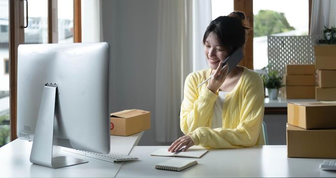 A portrait of Asian woman, e-commerce employee sitting in the office full of packages on the table using a laptop and smartphone, for SME business, e-commerce, technology and delivery business..