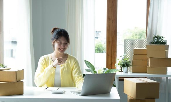 A portrait of Asian woman, e-commerce employee sitting in the office full of packages on the table using a laptop and calculator, for SME business, e-commerce, technology and delivery business..