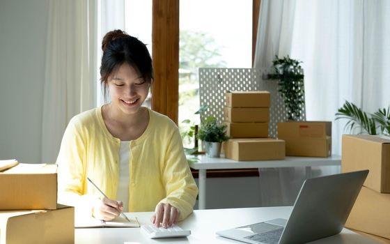 A portrait of Asian woman, e-commerce employee sitting in the office full of packages on the table using a calculator, for SME business, e-commerce, technology and delivery business..