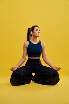 Front view of sporty girl sitting with bent legs, yoga doing indoors. Flexible brunette female looking up, holding hands, with mudras. Isolated on yellow studio background.