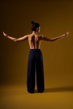 Back view of slim, flexible female practicing yoga at studio. Brunette girl with bare back standing, raising arms, stretching, meditating. Isolated on yellow studio background.
