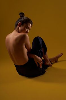 Back view of seductive girl with bare back sitting backwards on floor. Flexible brunette with hair bun doing yoga, looking down, breezing. Isolated on yellow studio background.