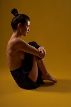 Side view of flexible girl sitting on floor, holding legs with arms. Pretty brunette with hair bun looking down, meditating, doing yoga. Isolated on yellow studio background.