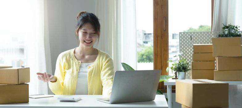 A portrait of Asian woman, e-commerce employee sitting in the office full of packages on the table using a laptop and calculator, for SME business, e-commerce, technology and delivery business..