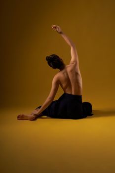 Back view of flexible girl with bare back sitting backwards on floor, with raised hand. Brunette with hair bun stretching, meditating. Concept of new age and yoga practicing.