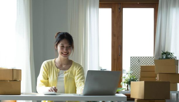 A portrait of Asian woman, e-commerce employee sitting in the office full of packages on the table using a laptop and calculator, for SME business, e-commerce, technology and delivery business..