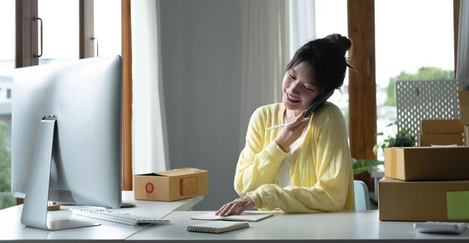 A portrait of Asian woman, e-commerce employee sitting in the office full of packages on the table using a laptop and smartphone, for SME business, e-commerce, technology and delivery business..