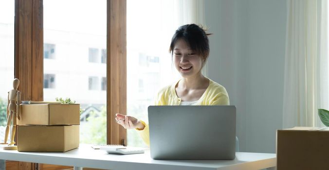 A portrait of Asian woman, e-commerce employee sitting in the office full of packages on the table using a laptop and calculator, for SME business, e-commerce, technology and delivery business..