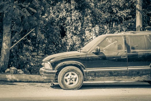 Old black and white picture of an old dirty car pick up truck at street road and cityscape with cars traffic palm trees bars and restaurants of Tulum in Mexico.