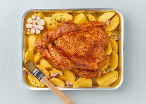 whole marinated chicken in sauce with raw potatoes, with brush in baking dish, top view