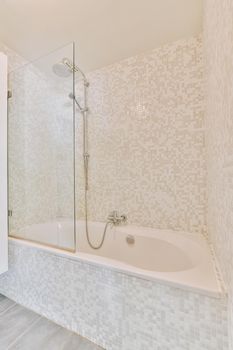 Bathtub with shower and glass partition placed in corner in small light bathroom with beige tile walls