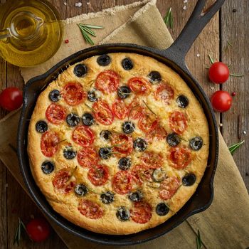 Focaccia, pizza in skillet, italian flat bread with tomatoes, olives and a rosemary. Wooden table