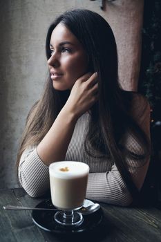 Beautiful girl sits in cafe in Christmas holidays, in background lights of garland. Brunette woman with long hair drinks cappuccino coffee, latte and looks out window, vertical, dark backdrop