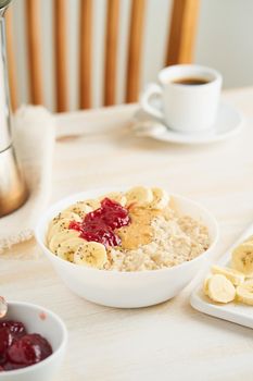 Oatmeal porridge, healthy vegan diet breakfast with strawberry jam, peanut butter, banana, chia on white wooden light background. Vertical.