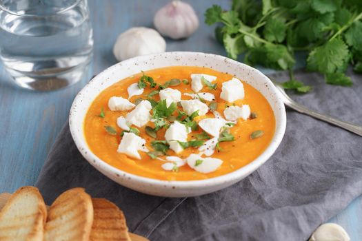 Pupmkin cream soup puree with ricotta, dietary vegetarian lunch on blue wooden table, side view. Glass of water, toast, parsley, garlic.