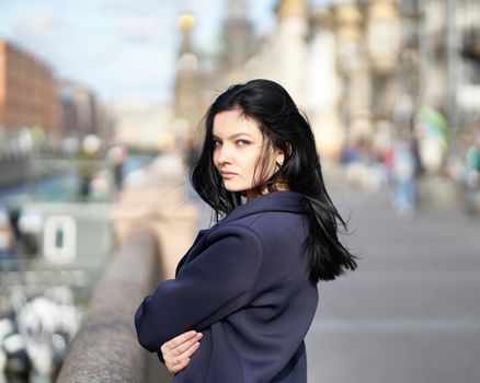 Portrait f beautiful intelligent brunette who walks down street of Saint-Petersburg in city center near bridge. Charming thoughtful woman with a long dark hair wanders alone, immersed in thoughts