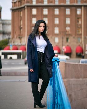 Charming thoughtful fashionably dressed woman with long dark hair travels through Europe, standing in city center of St. Petersburg. A beautiful girl wanders alone through autumn streets, vertical