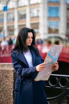 Young woman standing on waterfront of big city and looking at guide, a tourist looking for for attractions. A charming thoughtful fashionably dressed woman with long dark hair travels through Europe